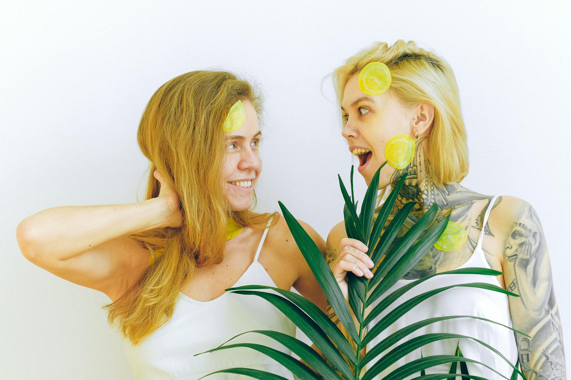 Duas mulheres sorrindo com fatias de limão e plantas tropicais, expressando alegria e diversão em ambiente interno
